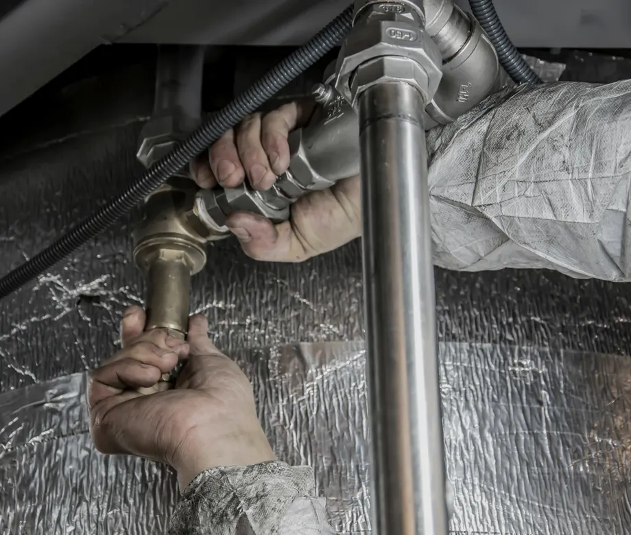 A plumber's two hands tightening a brass union on a chrome pipe under a sink, against foil-wrapped insulation.